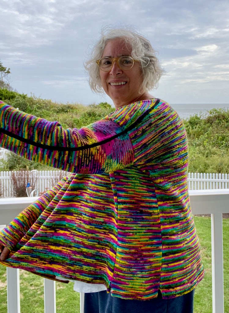 Modeled side view of black and neon pullover with front held out in front of body to show the construction of the garment