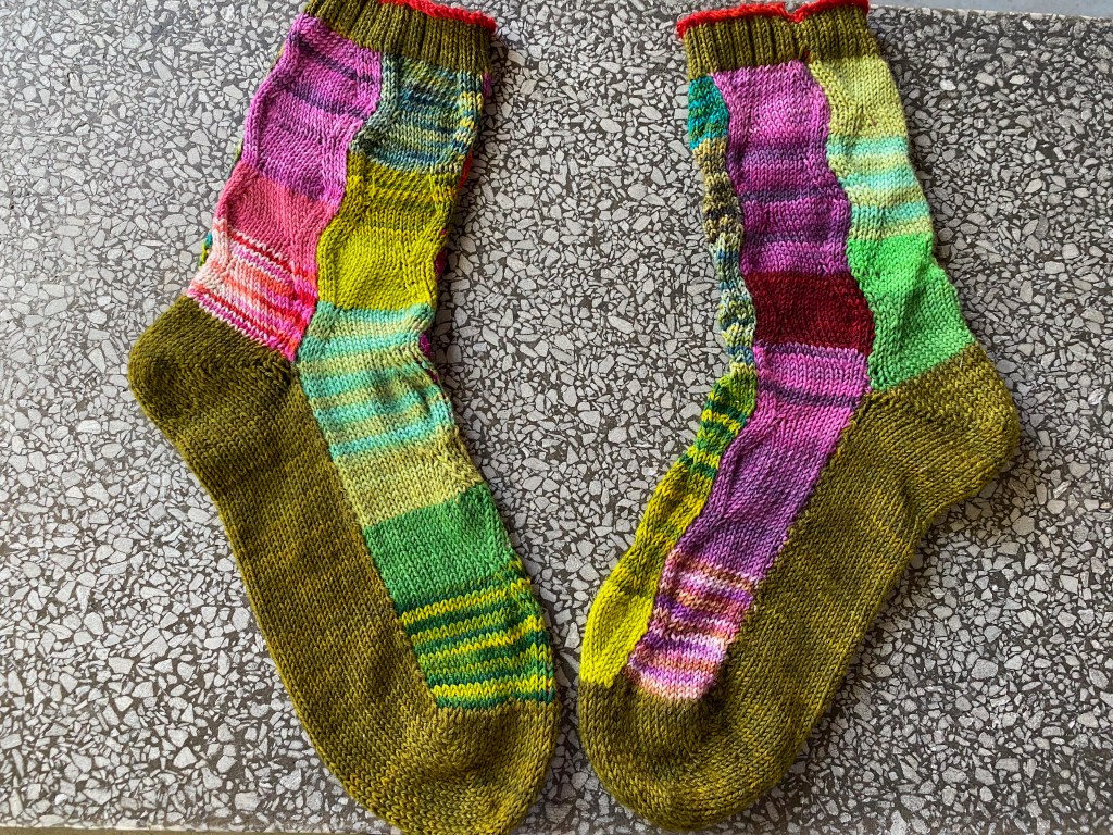 Two socks machine-knitted in wavy panels shaped with increases and decreases, using many different yarns in pinks and greens