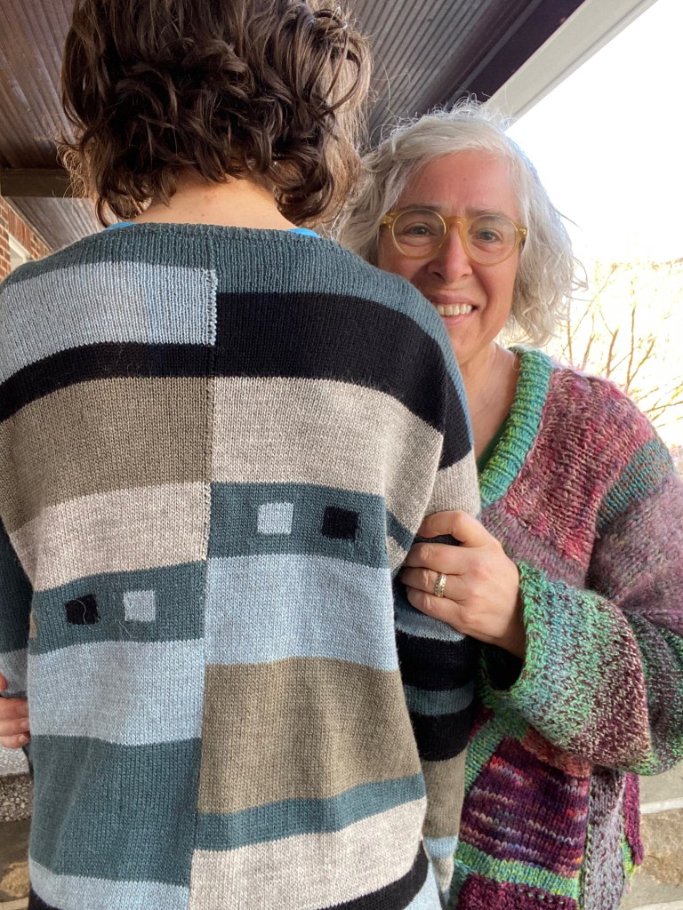 Mother-daughter sweaters of hand-knit patchwork pullover and machine-knit paneled pullover with intarsia and embroidery. Back view of machine-knit pullover