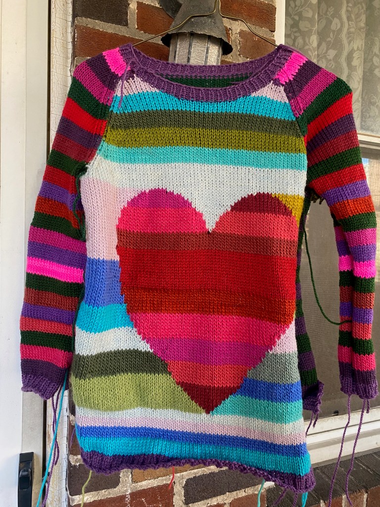 Machine knit intarsia heart in reds against background in cool colors, front of sweater